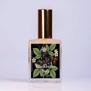 May include: A clear glass perfume bottle with a gold cap and a rectangular label. The label features a black background with a botanical illustration of green leaves, white flowers, and dark blackberries, with the text Blackberry Noir PERFUME.