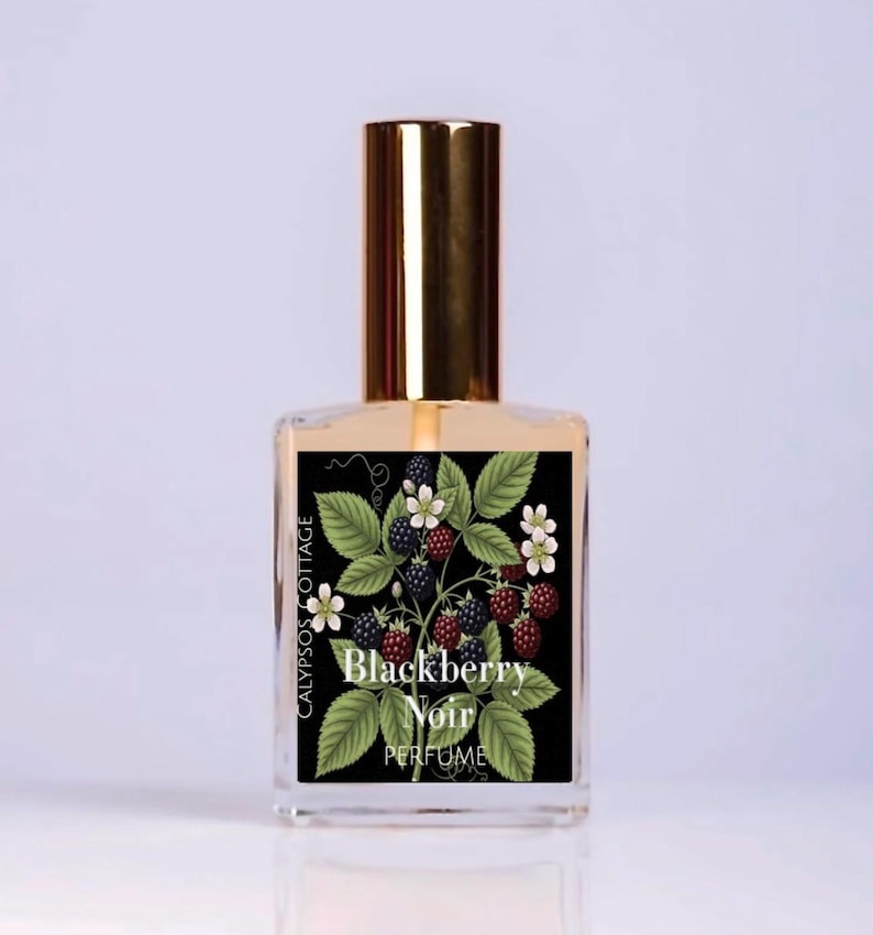 May include: A clear glass perfume bottle with a gold cap and a rectangular label. The label features a black background with a botanical illustration of green leaves, white flowers, and dark blackberries, with the text Blackberry Noir PERFUME.