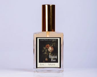 May include: A clear glass perfume bottle with a gold cap. The label features a floral painting and the words "Turkish Rose." The bottle is labeled "Calypso's Cottage" and contains 5 ml of perfume.