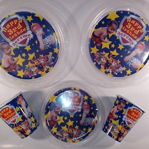 Op de afbeelding: Drie doorzichtige plastic borden en twee papieren bekers met een blauwe achtergrond met gele sterren, stripfiguren van honden en een schild met de tekst Happy 3rd Birthday Jerome, inclusief een foto van een jong kind.