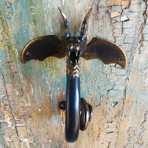 May include: A black metal dragon door knocker with gold accents. The dragon has wings and a long tail. The knocker is mounted on a wooden door.