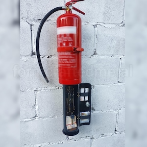Peut inclure: Un extincteur rouge transformé en unité de rangement murale. L'extincteur conserve sa poignée et son tuyau d'origine. Un compartiment noir en dessous contient des clés, de l'argent et d'autres petits objets. L'unité est fixée sur un mur de briques blanches.