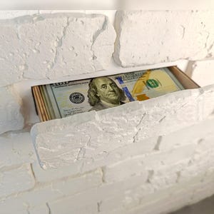 Puede incluir: Un fajo de billetes de cien dólares estadounidenses es parcialmente visible dentro de un compartimento oculto en una pared de ladrillos blancos. Los billetes presentan un retrato de Benjamin Franklin.
