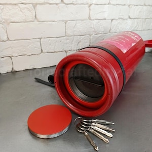 Puede incluir: Una caja fuerte cilíndrica de metal rojo diseñada para parecer un extintor. La tapa superior está retirada, revelando una abertura circular con un interior roscado. Cuatro llaves plateadas están dispuestas en abanico junto a la tapa roja.