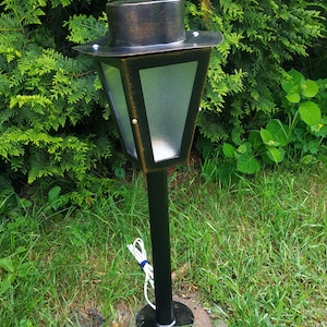 May include: A black outdoor lamp post with a bronze-colored top and trim. The lamp has a clear glass panel and is set in a grassy area with green foliage in the background.