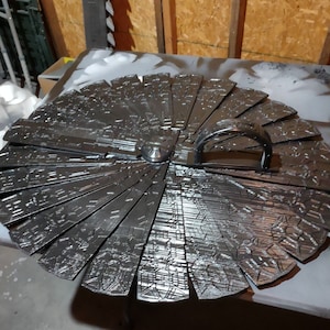 Könnte beinhalten: Ein großes, silberfarbenes, kreisförmiges Objekt mit einem Griff. Das Objekt besteht aus vielen rechteckigen Segmenten, die jeweils ein detailliertes, strukturiertes Muster aufweisen. Der Griff ist ein gebogener Metallbügel.