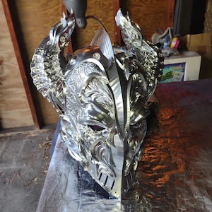 May include: A highly detailed, silver-colored fantasy helmet with intricate floral and dragon-like designs. The helmet features a visor, cheek guards, and prominent horns, suggesting a medieval or mythical theme. The image includes the text "3D ARTGUY" at the bottom.