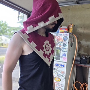 Può includere: Un cappuccio bordeaux con la parte superiore a punta e un bordo color crema. Il cappuccio presenta un bavaglino bordeaux abbinato con un disegno di teschio e ingranaggio. La persona nell'immagine indossa una canotta nera.