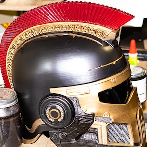 May include: A black and gold helmet with a red crest. The helmet features intricate gold detailing around the crest and face guard. A circular emblem with a laurel wreath is on the side. The helmet is likely a costume or decorative item.