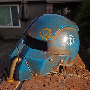 Pode incluir: Um capacete azul desgastado com aparência enferrujada, apresentando um emblema dourado de engrenagem e um símbolo branco. O capacete tem uma viseira e um tubo lateral. O texto "Vauls_Anvil" é visível.