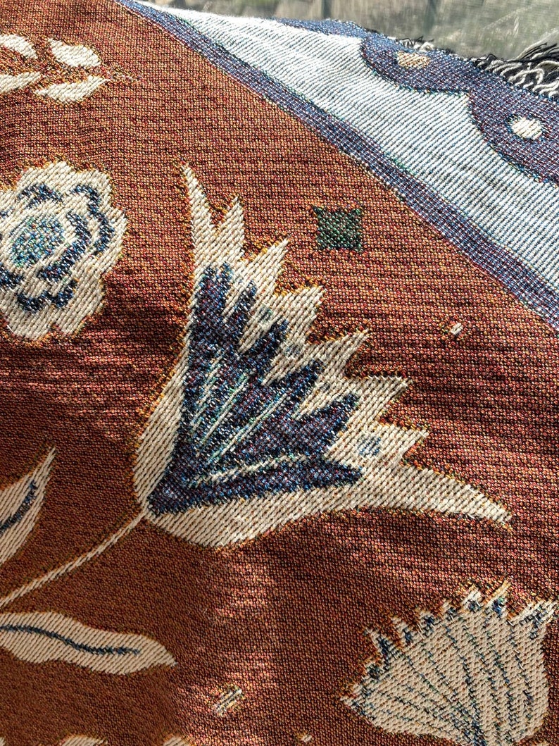 Op de afbeelding: Close-up van een bruine stof met een geweven ontwerp met witte en blauwe bloemenpatronen. De stof heeft een textuur en een subtiele glans.