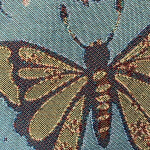 Op de afbeelding: Een close-up van een geweven wandkleed met een grote vlinder met bruine, gele en blauwe vleugels op een blauwe achtergrond.
