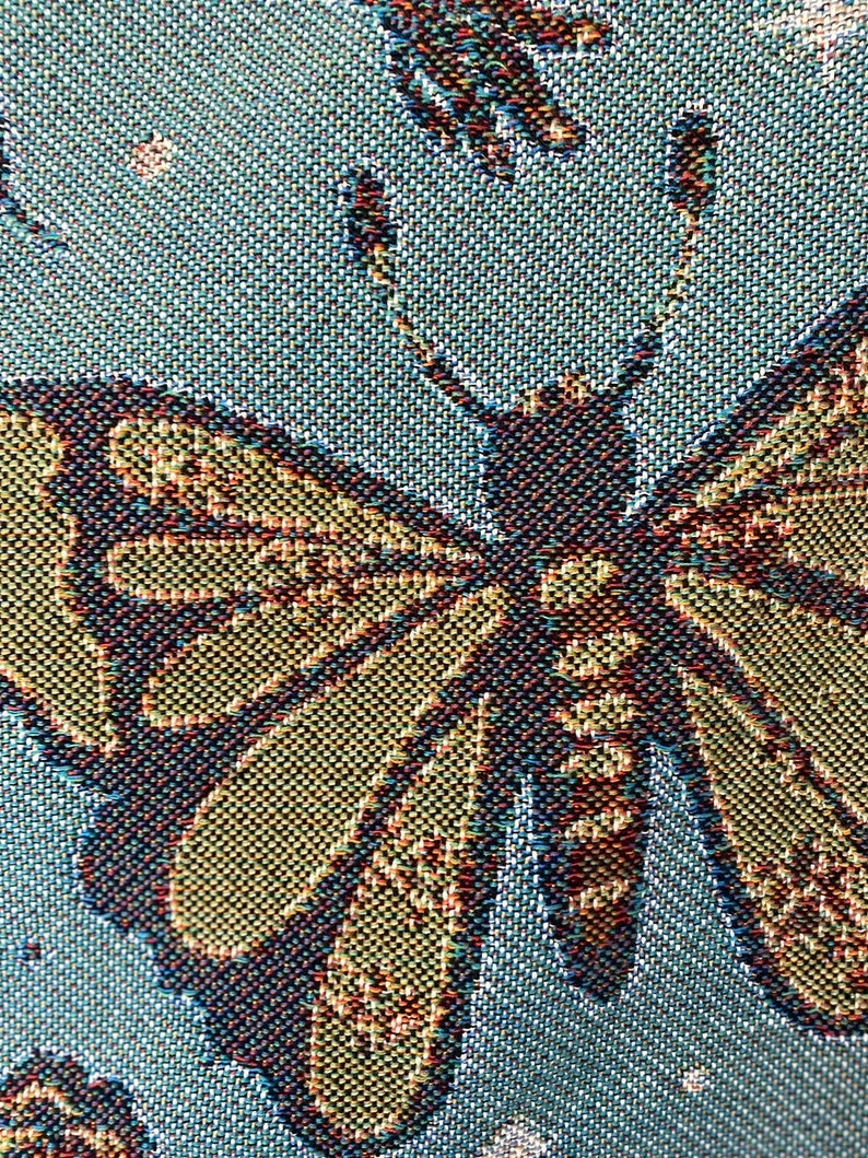 Op de afbeelding: Een close-up van een geweven wandkleed met een grote vlinder met bruine, gele en blauwe vleugels op een blauwe achtergrond.