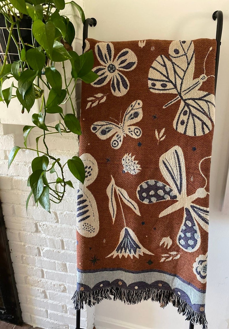 Op de afbeelding: Een bruin geweven deken met een franje rand heeft een patroon van witte vlinders en bloemen op een bruine achtergrond.