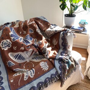 Op de afbeelding: Een bruin geweven deken met een blauwe rand en een wit en blauw vlinderpatroon. De deken is over een witte fauteuil met een franje rand gelegd.