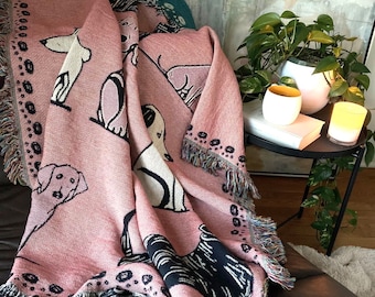 Könnte beinhalten: Eine rosa gewebte Wolldecke mit einem schwarz-weißen Hundemuster. Die Decke hat an allen Seiten Fransen und ist über einen braunen Ledersessel gelegt.