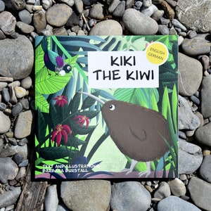 Könnte beinhalten: Ein Kinderbuch mit dem Titel "Kiki the Kiwi" mit Illustrationen eines Kiwis, tropischer Pflanzen und eines Vogels. Das Buch liegt auf einem Hintergrund aus grauen und braunen Flusssteinen.