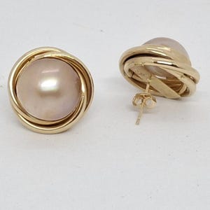 Peut inclure: Une paire de boucles d'oreilles dorées. Chaque boucle d'oreille présente une perle rose clair entourée d'un motif en métal doré torsadé. Les boucles d'oreilles sont posées sur un fond blanc.