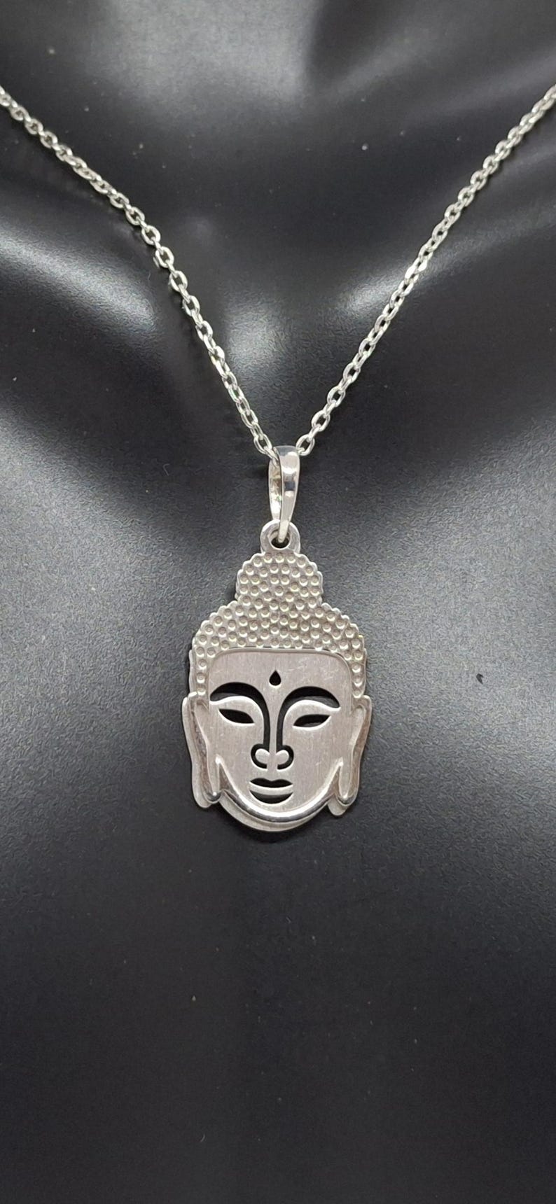 Puede incluir: Un colgante de tono plateado con forma de cabeza de Buda estilizada, con una parte superior texturizada y rasgos faciales suaves, suspendido de una delicada cadena plateada. El colgante presenta ojos recortados y un peque&ntilde;o punto en la frente.