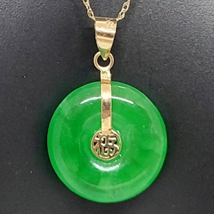 May include: A gold-coloured chain necklace with a bright green jade pendant. The pendant is a circular disc with a gold-coloured accent piece featuring Chinese characters. The necklace is displayed against a black background.