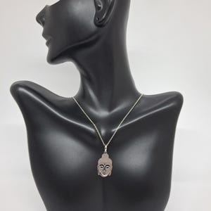 Puede incluir: Un collar de tono plateado con un colgante en forma de cabeza de Buda estilizada. El colgante tiene una superficie texturizada en el cabello y rasgos faciales suaves, suspendido de una delicada cadena plateada con eslabones est&aacute;ndar.