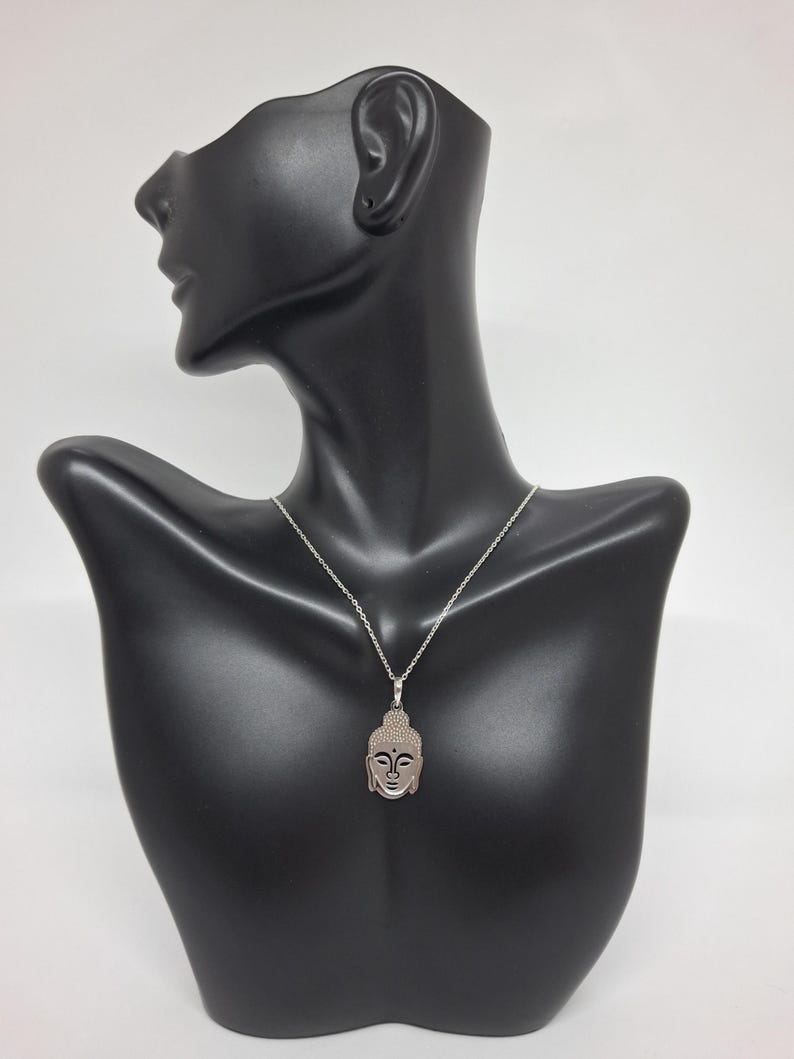 Puede incluir: Un collar de tono plateado con un colgante en forma de cabeza de Buda estilizada. El colgante tiene una superficie texturizada en el cabello y rasgos faciales suaves, suspendido de una delicada cadena plateada con eslabones est&aacute;ndar.
