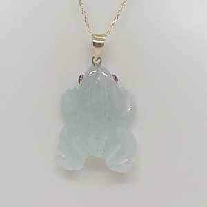 Puede incluir: Un collar de cadena de oro con un colgante de rana de jade. La rana tiene ojos rojos y está tallada en un estilo realista y detallado.