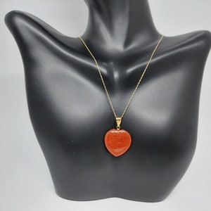 Puede incluir: Un collar de cadena de oro con un colgante de corazón rojo. El colgante está hecho de una piedra roja con una textura rugosa.