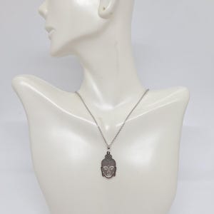 Puede incluir: Un collar de tono plateado con un colgante en forma de cabeza de Buda estilizada. El colgante tiene un acabado met&aacute;lico liso con detalles grabados que delinean los rasgos faciales y el cabello. Cuelga de una cadena fina de tono plateado.