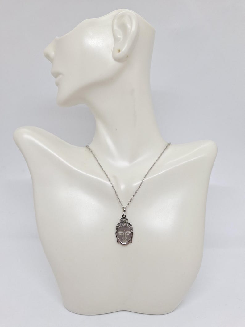 Puede incluir: Un collar de tono plateado con un colgante en forma de cabeza de Buda estilizada. El colgante tiene un acabado met&aacute;lico liso con detalles grabados que delinean los rasgos faciales y el cabello. Cuelga de una cadena fina de tono plateado.
