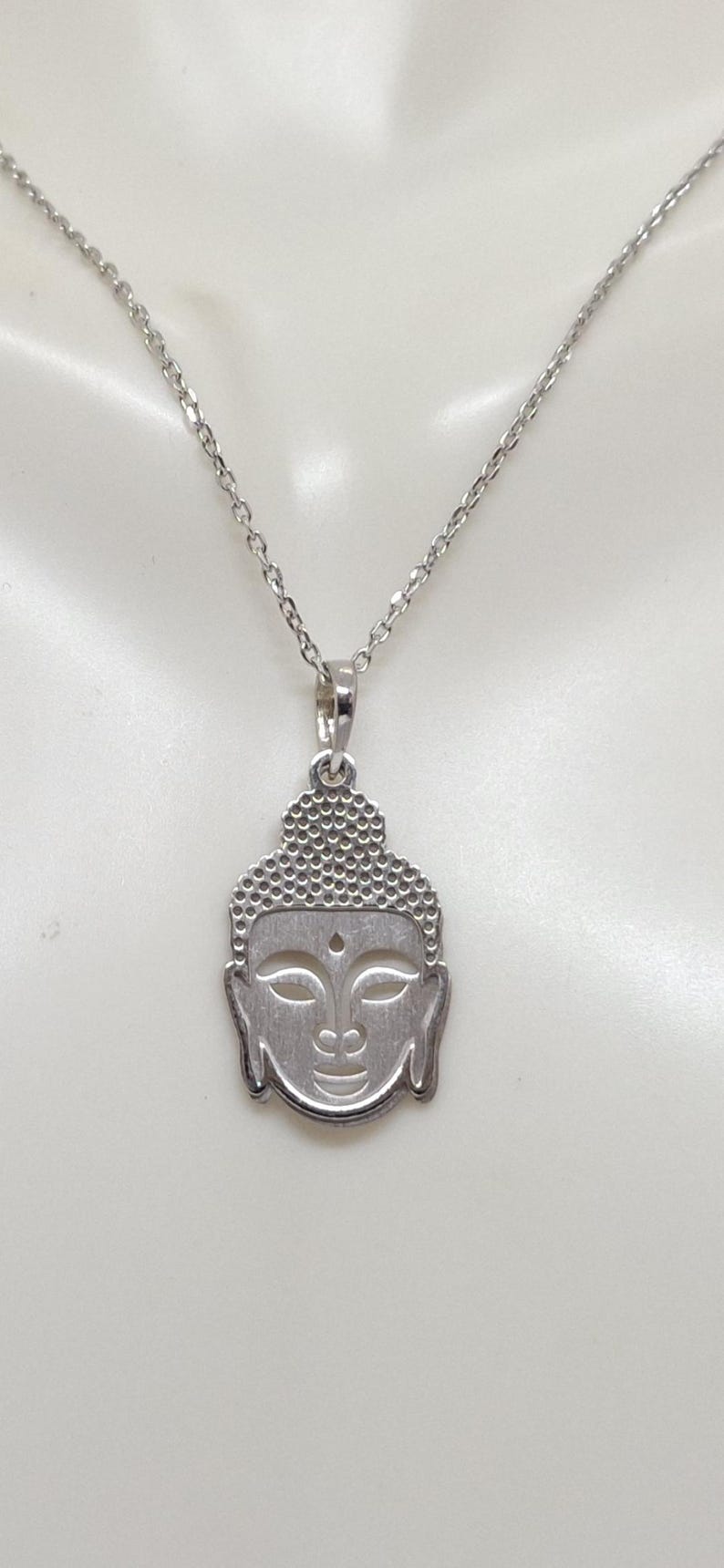 Puede incluir: Un collar de tono plateado con un colgante en forma de cabeza de Buda. El colgante presenta un patr&oacute;n texturizado de puntos en la parte superior que representa el cabello, con detalles lisos recortados para los ojos y un peque&ntilde;o punto en la frente.