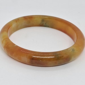 Puede incluir: Un brazalete de jade redondo en tonos naranja y amarillo. La piedra pulida tiene una superficie lisa y un aspecto natural. El brazalete es una joya.