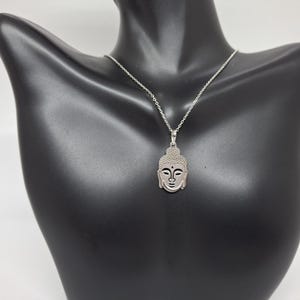 Puede incluir: Un collar de tono plateado con un colgante en forma de cabeza de Buda estilizada. El colgante tiene una secci&oacute;n superior texturizada y rasgos faciales suaves, suspendido de una delicada cadena plateada con un dise&ntilde;o de eslabones est&aacute;ndar.