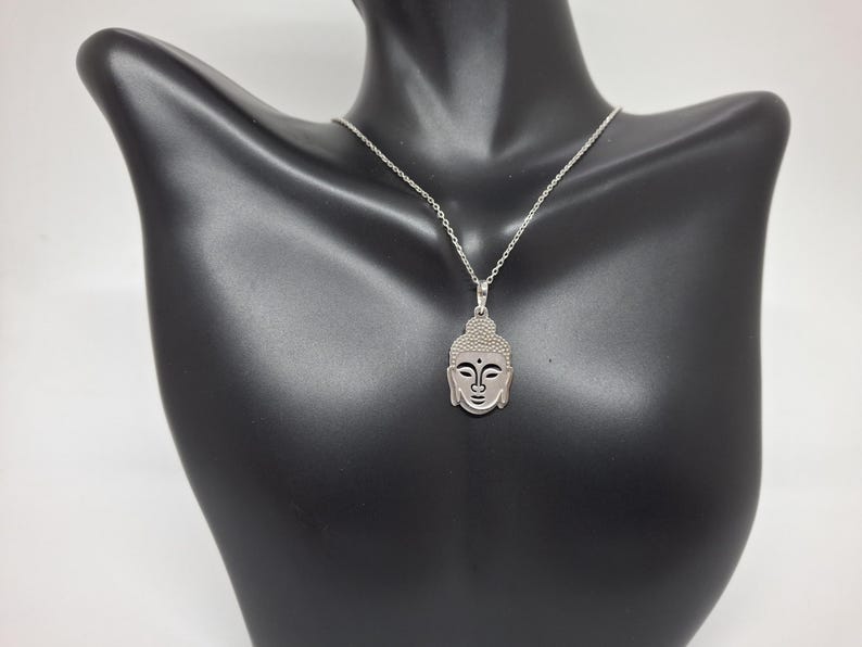 Puede incluir: Un collar de tono plateado con un colgante en forma de cabeza de Buda estilizada. El colgante tiene una secci&oacute;n superior texturizada y rasgos faciales suaves, suspendido de una delicada cadena plateada con un dise&ntilde;o de eslabones est&aacute;ndar.