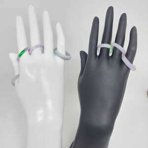 K&ouml;nnte beinhalten: Drei Jade-Ringe, einer gr&uuml;n und zwei lavendelfarben, werden auf zwei Schaufensterpuppenh&auml;nden pr&auml;sentiert. Die H&auml;nde sind wei&szlig; und schwarz.