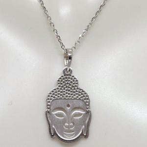Puede incluir: Un collar de tono plateado con un colgante en forma de cabeza de Buda. El colgante presenta un patr&oacute;n texturizado de puntos en la parte superior que representa el cabello, con detalles lisos recortados para los ojos y un peque&ntilde;o punto en la frente.