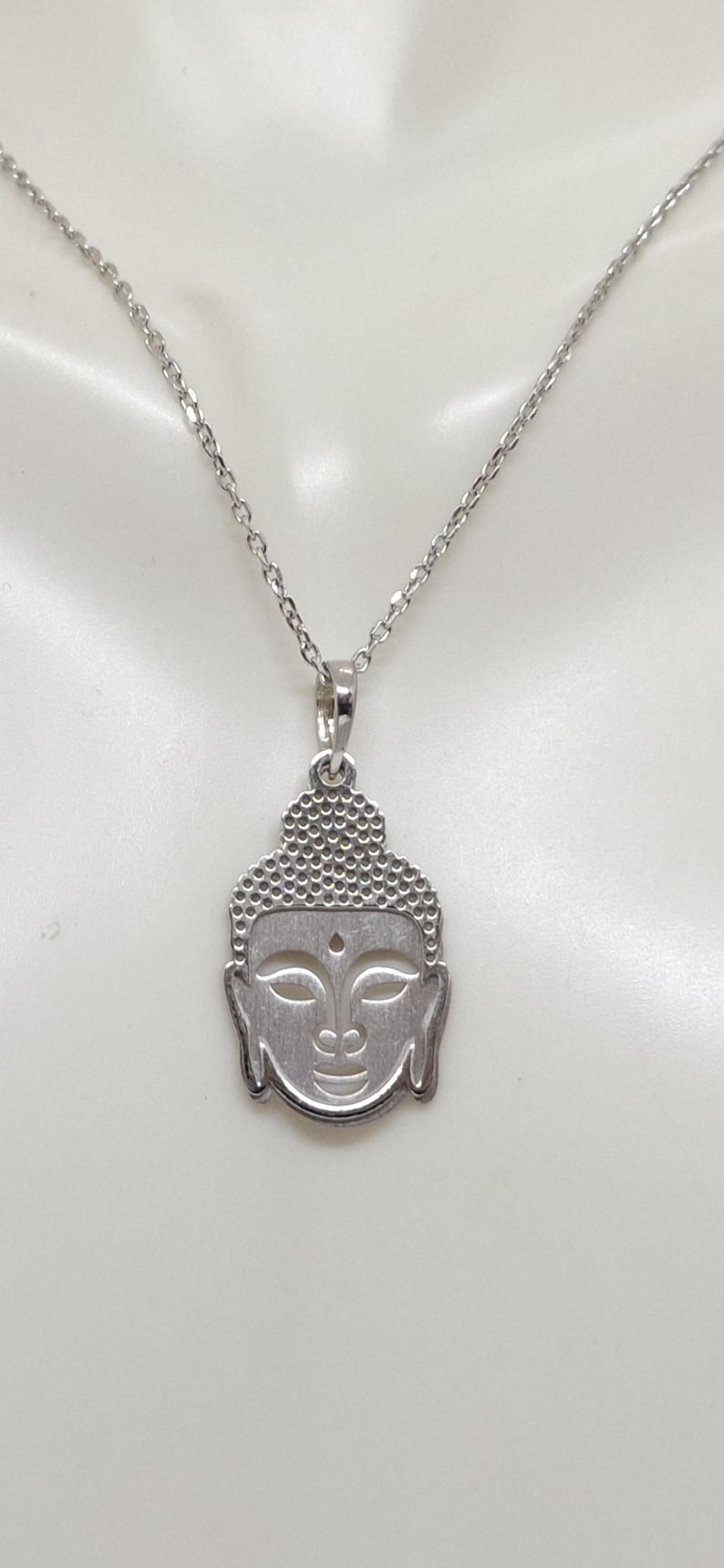 Puede incluir: Un collar de tono plateado con un colgante en forma de cabeza de Buda. El colgante presenta un patr&oacute;n texturizado de puntos en la parte superior que representa el cabello, con detalles lisos recortados para los ojos y un peque&ntilde;o punto en la frente.
