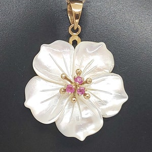 Puede incluir: Un collar de oro con un colgante de flor de nácar blanco de cinco pétalos. El centro de la flor está acentuado con pequeñas piedras preciosas redondas de color rosa engastadas en oro, con finos estambres dorados que se extienden.