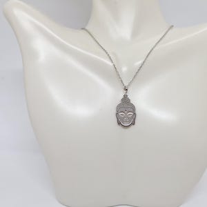 Puede incluir: Un collar de tono plateado con un colgante en forma de cabeza de Buda. El colgante presenta un patr&oacute;n texturizado de puntos en el cabello y rasgos faciales suaves y definidos. Cuelga de una cadena fina de metal de tono plateado.