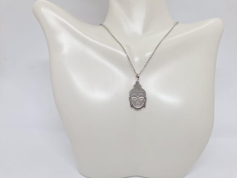Puede incluir: Un collar de tono plateado con un colgante en forma de cabeza de Buda. El colgante presenta un patr&oacute;n texturizado de puntos en el cabello y rasgos faciales suaves y definidos. Cuelga de una cadena fina de metal de tono plateado.