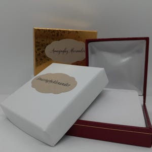 Puede incluir: Un joyero blanco con una etiqueta festoneada de color canela que dice AmazingbyAlexander, colocado junto a un joyero granate abierto con interior blanco y una caja dorada con una etiqueta de color canela a juego en el fondo.