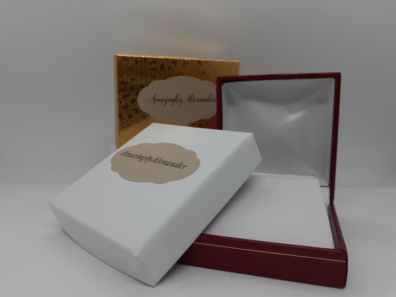 Puede incluir: Un joyero blanco con una etiqueta festoneada de color canela que dice AmazingbyAlexander, colocado junto a un joyero granate abierto con interior blanco y una caja dorada con una etiqueta de color canela a juego en el fondo.