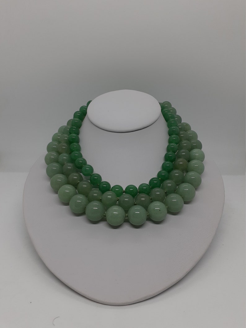 May include: A three-strand necklace made of light green jade beads. The beads are round and smooth, and the necklace is displayed on a white mannequin bust.
