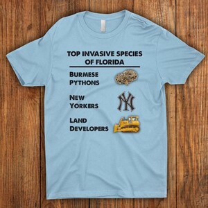 Könnte beinhalten: Hellblaues T-Shirt mit dem Text "TOP INVASIVE SPECIES OF FLORIDA". Das Shirt listet burmesische Pythons, New Yorker und Landentwickler mit entsprechenden Grafiken auf. Das Shirt besteht aus einem weichen Material.