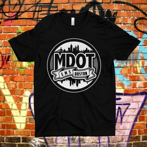 Può includere: Maglietta nera con un grafico circolare bianco che presenta uno skyline e il testo "MDOT EMS BOSTON".
