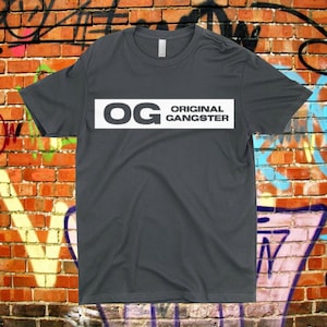 May include: A dark gray t-shirt with a white rectangle on the front. The rectangle has the text "OG ORIGINAL GANGSTER" in white.