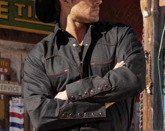 May include: A black cowboy hat and sunglasses are worn with a black button-down shirt and jeans. The shirt has red stitching and accents. A belt with a large silver buckle is visible. The image includes the "Stars & Stripes" logo.