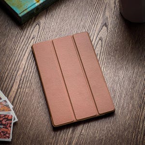 Peut inclure: Un étui folio en cuir marron avec trois panneaux. L'étui est fermé et posé à plat sur une surface en bois.