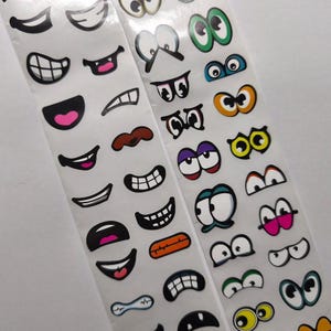 Op de afbeelding: Twee stroken stickers in cartoonstijl met verschillende mond- en oogontwerpen. De monden hebben verschillende uitdrukkingen, waaronder glimlachen en fronsen, met roze en witte accenten. De ogen hebben verschillende kleuren en vormen, sommige met meerdere pupillen.