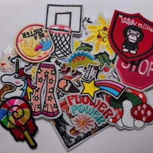 Puede incluir: Una colección de parches bordados con varios diseños, incluyendo una canasta de baloncesto, un girasol, un unicornio, una piruleta y las palabras "Flower Power". Los parches presentan colores brillantes y costuras detalladas.